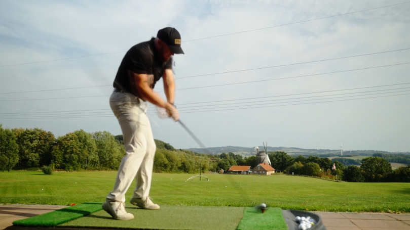 Eine Person beim Golfspielen auf einer offenen Wiese mit grünen Hügeln im Hintergrund.