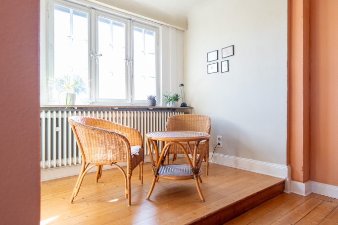 Ein heller Raum mit zwei Korbstühlen und einem Tisch, umgeben von Fenstern.