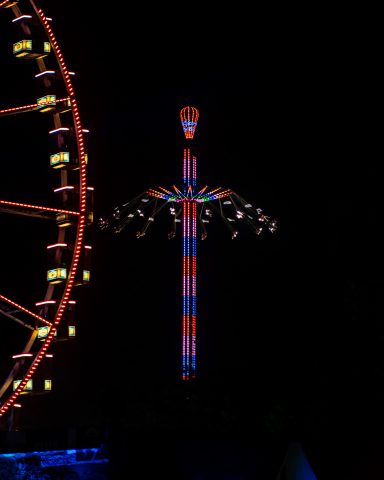 Ein Riesenrad und ein beleuchteter Aussichtsturm bei Nacht, beide in bunten Lichtern.