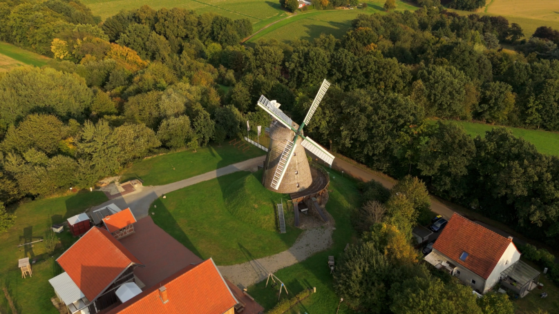Luftaufnahme einer Windmühle umgeben von Wiesen und Bäumen, mit nahegelegenen Häusern.