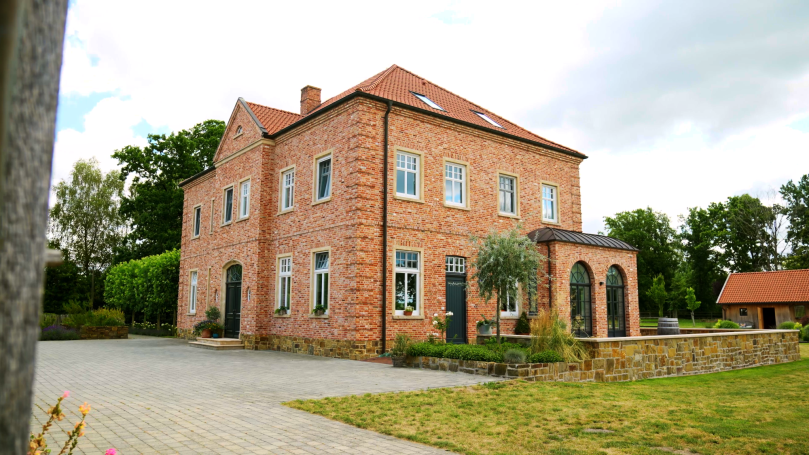 Rotes Backsteinhaus mit Garten und Fensterfront, umgeben von grünen Bäumen.