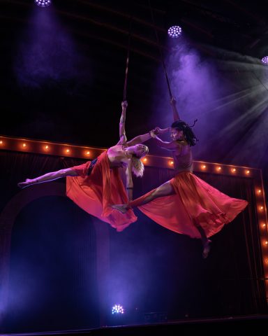 Zwei Akrobatinnen in roten Kleidern schwingen an Stoffbändern über einer Bühnenbeleuchtung.