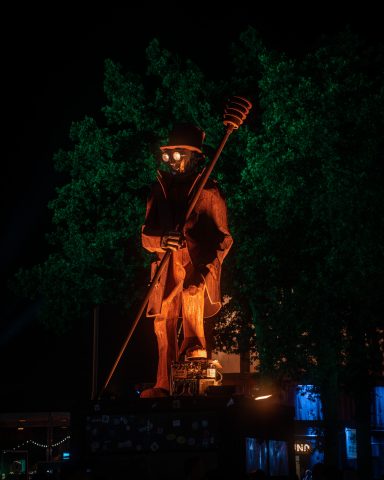 Große statue eines wandernden Mannes mit Stab, umgeben von grünen Bäumen und Nachtbeleuchtung.