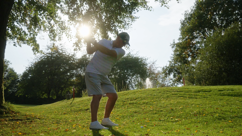 Mann schlägt Golfball auf einer grünen Wiese im Sonnenschein.