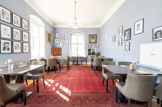 Heller Raum mit Tischen, Stühlen und großen Fenstern, Wänden voller Bilder.