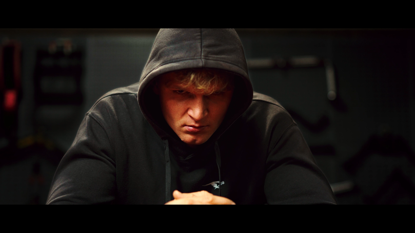 Ein muskulöser Mann in einem schwarzen Kapuzenpullover, nachdenklich am Tisch sitzend.