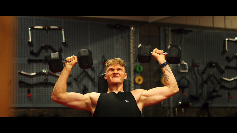 Mann hebt Gewichte im Fitnessstudio, lächelt dabei.