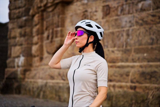 Frau in Radkleidung und Helm, die eine Sonnenbrille anpasst, vor einer steinernen Wand.
