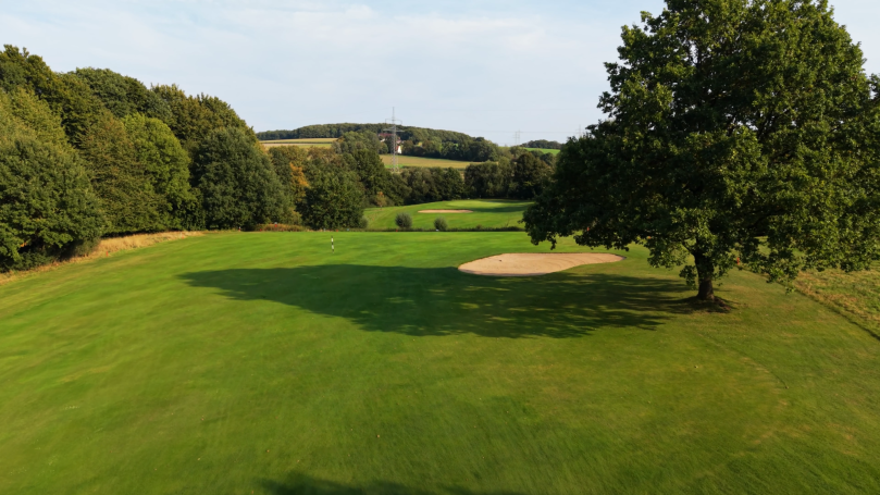 Weitläufige grüne Landschaft mit Bäumen und einem Sandbunker auf einem Golfplatz.